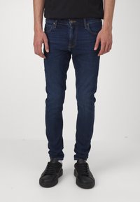 Tmavě modré skinny džíny s klasickým designem pěti kapes, vyrobené z denimu. Nosené s černými šněrovacími teniskami. Jednoduché detaily a švy.