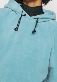Persona che indossa un felpa leggera di colore blu chiaro con cordini neri regolabili sopra un top nero lavorato a collo alto.