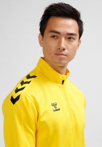 Gul sportjacka med hög krage, svart logotyp och accenter, med dragkedja och framträdande svarta chevronmönster på ärmarna.