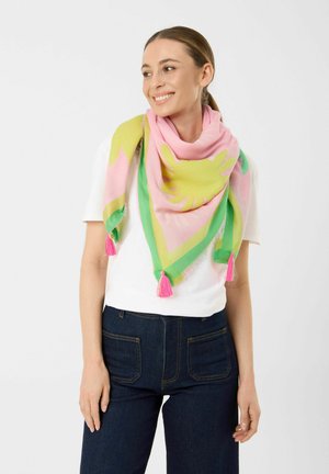 Woman smiling, wearing white short-sleeve shirt, dark blue jeans, and a colorful pink, green, and yellow scarf with tassels around her neck.