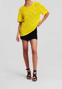 Top amarillo de manga corta tejido con un patrón texturizado, combinado con pantalones cortos negros con textura y sandalias negras de tacón alto con tiras.