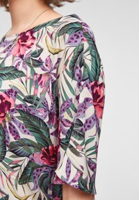 Blusa floreale con maniche a campana, caratterizzata da una stampa tropicale colorata in rosa, viola e verde su uno sfondo chiaro, tessuto strutturato.