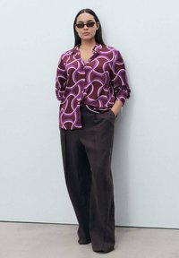 Camicia a maniche lunghe con bottoni di un profondo bordeaux con motivo a onde viola; abbinata a pantaloni larghi scuri. Modello con occhiali da sole neri.