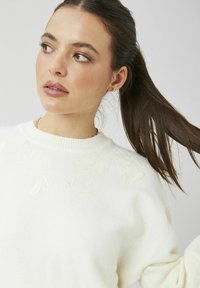 Jeune femme aux cheveux bruns attachés en queue de cheval, portant un pull blanc avec une broderie florale discrète, regardant vers sa gauche.