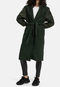 Žena v tmavě zeleném kabátu s páskem a rukávy z různých materiálů, bílou košili, černých skinny džínách a černých teniskách, postávající s rukama na bok.