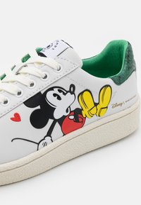 Vit sneaker med Mickey Mouse-grafik, gula handskar, röda shorts, grön inredning, vita snören och strukturerad sula. Disney-logotyp på sidan.