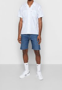 Camisa blanca de botones de manga corta con rayas verticales azules, combinada con pantalones cortos de mezclilla azul y zapatillas deportivas blancas, con calcetines blancos.
