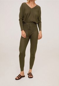 Olivově zelený dvoudílný outfit s volným topem s výstřihem do V a přiléhavými kalhotami s pasem na šňůrku, doplněný hnědými šlapkami.