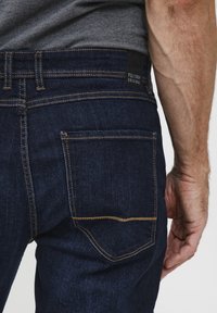 Jeans de mezclilla azul oscuro con costuras doradas en contraste, bolsillos estándar y una etiqueta negra en la cintura. Muestra una vista cercana de la parte trasera.