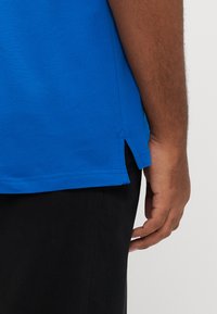 Blå poloshirt med et tekstureret stof, med en sidelomme i kanten, kombineret med sorte bukser. Nærbillede af skjortens bagside og hånd.