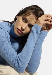 Jasnoniebieski, prążkowany sweter z golfem, wykonany z miękkiego materiału, dopasowany krój, z długimi rękawami opierającymi się na skrzyżowanych ramionach przy brodzie.