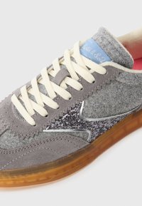 Sneaker in suede grigio con accenti argento, dettaglio glitter, lacci color crema e etichetta blu. Suola arancione semi-trasparente. Materiali testurizzati.