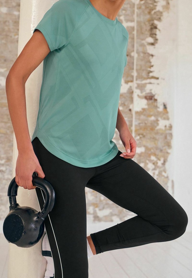 Personne portant un t-shirt à manches courtes couleur sarcelle et un legging noir, tenant un kettlebell et s'équilibrant sur une jambe contre un pilier blanc dans une pièce lumineuse.