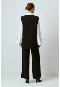 Black sleeveless vest over a white shirt, paired with high-waisted black pants featuring textured side accents, and pointed toe black boots.