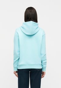 Γυναίκα με ίσια σκούρα μαλλιά που φοράει ανοιχτό μπλε φούτερ με κουκούλα και σκούρο μπλε τζιν, στέκεται γυρισμένη πλάτη σε απλό λευκό φόντο.