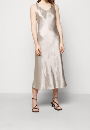 Woman wearing a sleeveless, metallic silver midi slip dress with black strappy heeled sandals, standing against a plain light background.