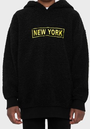 Person iført en sort, fluffy hættetrøje med "NEW YORK" broderet i gult på brystet, synlig fra nakke til hofter.