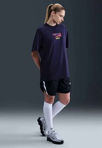 Sieviete stāv tērpusies tumši zilā Barca T-kreklā, melnās šortos ar baltām svītrām, baltās ceļgalu augstumā zeķēs un baltās Nike stādē. Fons ir tumši pelēks.