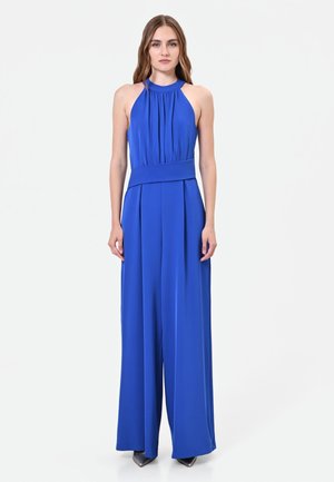 Donna che indossa una tuta senza maniche blu reale con collo all'americana, pantaloni larghi e scarpe nere con punta, in piedi davanti a uno sfondo uniforme.