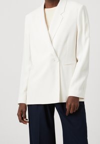 Blazer blanco a medida con cierre de un solo botón, solapas de muesca y hombros estructurados. Tejido suave, corte relajado y longitud ligeramente mayor.