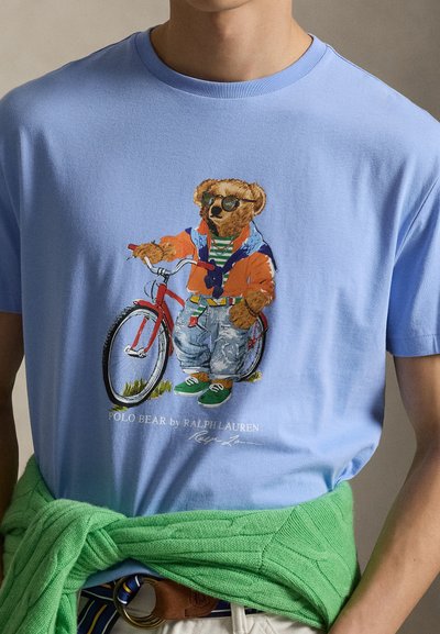 Μπλούζα με γραφικό ενός αρκούδου που φοράει γυαλιά ηλίου, πολύχρωμο μπουφάν, τζιν και πράσινα αθλητικά, που στέκεται δίπλα σε ένα κόκκινο ποδήλατο.