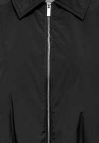 Κοντινή λήψη στο μπροστινό μέρος ενός μαύρου σακακιού με ασημένιο μεταλλικό φερμουάρ που τρέχει κάθετα κάτω από μια διπλωμένη γιακά.