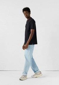 Polo nero con colletto e bottoni, abbinato a jeans azzurri chiari. Il modello indossa sneakers grigie, in piedi di fronte a uno sfondo neutro.