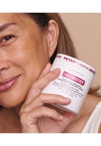 Conteneur blanc étiqueté "Encore plus doux" avec du texte rouge, présentant une surface mate lisse. Détails visibles du couvercle du produit et impression de prise sur le côté.