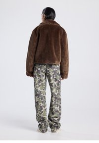 Ruskea keinoturkistakki, jossa korkea kaulus, yhdistetty harmaisiin ja vihreisiin maastokuvioituihin housuihin. Takissa on rento istuvuus ja sileä pinta.