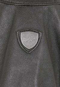 Aufnäher aus schwarzem Leder mit einem metallischen Schild, auf dem "Mauritius" in kursiver Schrift eingraviert ist. Strukturierte Oberfläche und detaillierte Nähte sind vorhanden.