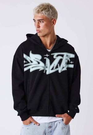 Jeune homme aux cheveux blonds courts portant un sweat à capuche noir zippé avec un grand texte blanc de style graffiti sur la poitrine.