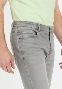 Graue Jeans aus Denim mit Slim Fit, ausgestattet mit fünf Taschen, dezenten Steppdetails und einem Reißverschluss, auf einem Mannequin präsentiert.
