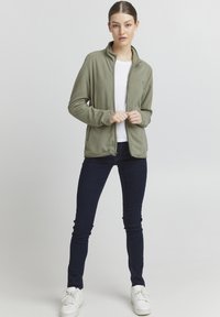 Fleecejakke i olivengrønn med høy krage, glidelås foran og sidelommer, kombinert med en hvit t-skjorte og mørke jeans.