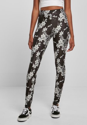 Svart leggings med et hvitt blomsterdesign, høyt liv, strekkbart stoff og en glatt tekstur. Sammen med svarte joggesko.