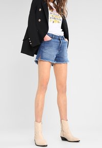 Denimshortsit, joiden helmassa on revitty reuna, yhdistetty valkoiseen graafiseen t-paitaan ja mustaan bleiseriin. Asun täydentävät beige cowboy-kengät.