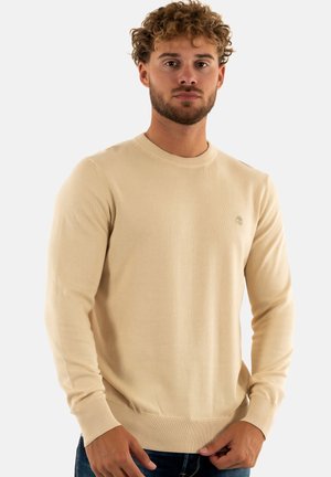 Maglione beige a maglia con colletto rotondo, polsini e orlo a coste, e un piccolo logo sul petto. Texture morbida e design aderente.