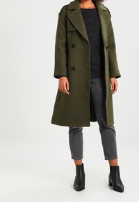 Olivgrön kappa i ullblandning med dubbelknäppning, överdimensionerad krage, svarta knappar, sidofickor, kombinerad med grå jeans och ankelboots.