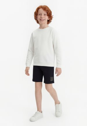 Garçon aux cheveux roux souriant, marchant, portant une chemise à manches longues blanche, un short noir avec un petit logo et des baskets blanches sur un fond blanc.