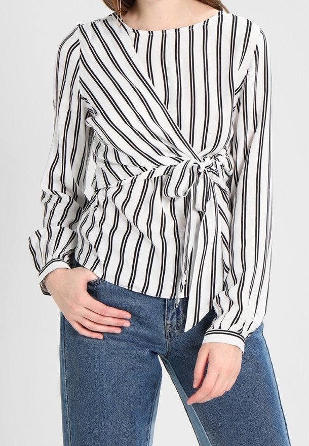 Femme portant un chemisier blanc à manches longues avec des rayures verticales noires et un nœud à l'avant, associé à un jean en denim bleu.