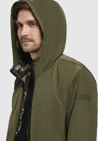 Olivegrüne Kapuzenjacke mit Reißverschluss, camouflagemuster Innenfutter und einem Patch am Ärmel. Glatte Textur, modernes Design.