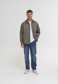 Veste gris clair avec un col, dotée de poches avant et d'une fermeture à boutons, associée à un jean bleu et des baskets blanches. T-shirt blanc uni en dessous.