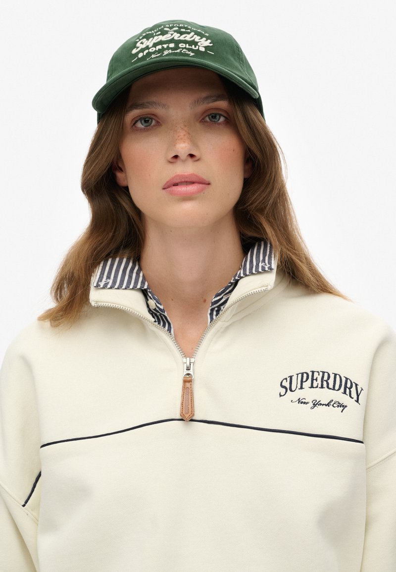 Crème kleurige sweatshirt met ritssluiting en marineblauwe accenten, voorzien van geborduurde "SUPERDRY" tekst. Heeft een gestreept overhemd eronder en een groene baseballpet.