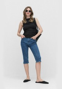 Svart ärmlös topp, blåcroppade denimjeans med framstygn, och svarta slip-on skor. Modellen poserar med händerna i fickorna och bär solglasögon.