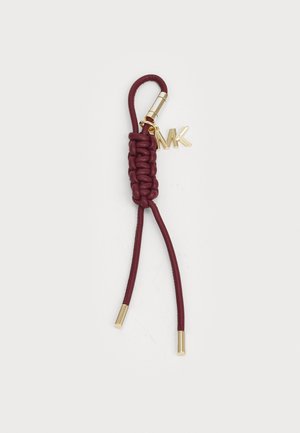 Porte-clés en cuir bordeaux avec un détail tressé, quincaillerie dorée, et un petit charm en forme de "MK" attaché. Deux lanières de cuir lisse s'étendent vers l'extérieur.