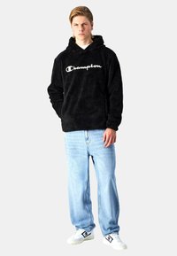 Sudadera negra de felpa con el logo blanco de "Champion", combinada con jeans rectos de color azul claro y zapatillas blancas. Ajuste y texturas casuales.