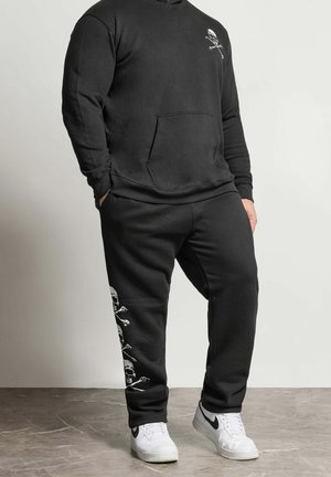 Hombre con sudadera negra con capucha y pantalones deportivos negros a juego con estampados blancos de calaveras y huesos cruzados, de pie sobre suelo gris, zapatillas blancas visibles.