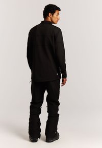 Schwarzer Pullover mit langen Ärmeln und hohem Kragen, kombiniert mit schwarzen lässigen Hosen und schwarzen Wanderschuhen, die ein dezentes Logo-Detail aufweisen.