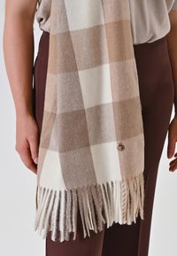 Beige en crème geruite sjaal gemaakt van zachte stof, met franjes aan de randen en een klein circulair accentdetail.