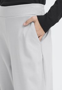 Un pantalon gris clair présente une texture lisse, une large ceinture et des poches latérales, avec une main reposant dans la poche gauche.