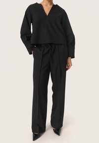 Svart kort topp med V-ringning och långa ärmar, ihop med svart högmidjade byxor med dragsko och veck. Svarta spetsiga klackar.